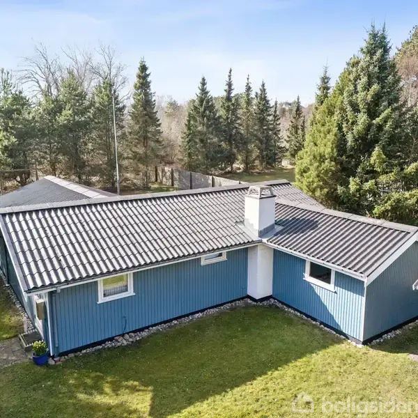 Blåt hus står stille omgivet af en have med grøn græsplæne. Omkring huset ses høje træer, som danner en skovagtig ramme.