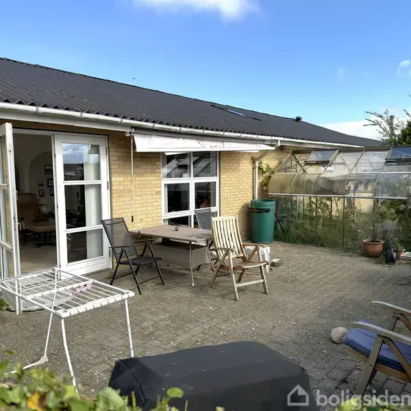 Et stenbelagt terrasseområde med bord og forskellige stole foran et gult murstenshus. Et lille drivhus står til højre, og en åben dobbeldør forbinder til en lys stue indendørs.