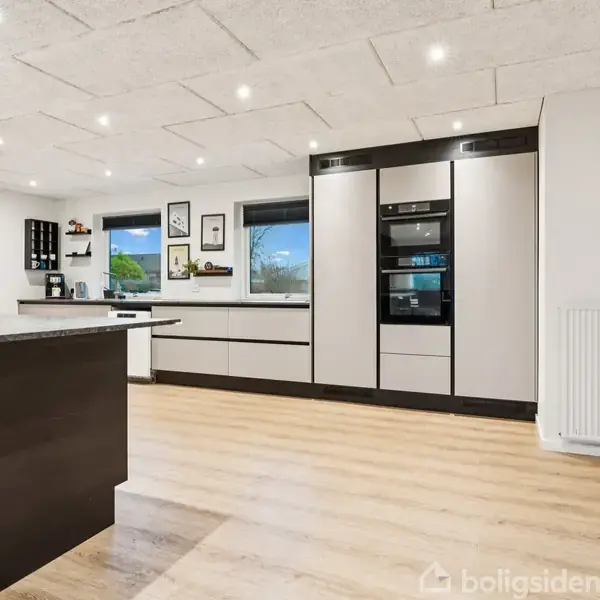 Køkken med højteknologiske hvidevarer og glatte, grå skabe. Handlepladerne er sorte, og rummet har trægulv. Store vinduer og lys overalt skaber en lys og moderne atmosfære.