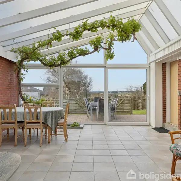 Et lyst vinterhaveværelse med gennemsigtigt tag og flisegulv. Et spisebord med stole står ved siden af en klatreplante, der dekorerer indvendigt. Udenfor er et terrassebord med udsigt til haven.