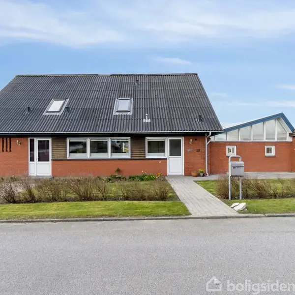 Et rødt murstenshus med sorte tagsten og flere vinduer står i en villakvarter. En grussti fører til hoveddøren, omgivet af en velplejet græsplæne og hække. Nummeret "62" er synligt ved hoveddøren.