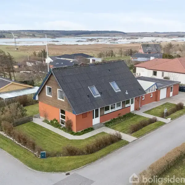 Et moderne hus med sort tag og røde mursten står på en hjørnegrund. Det har en velplejet have og er omgivet af en vej og nabohuse. Landskab i baggrunden.