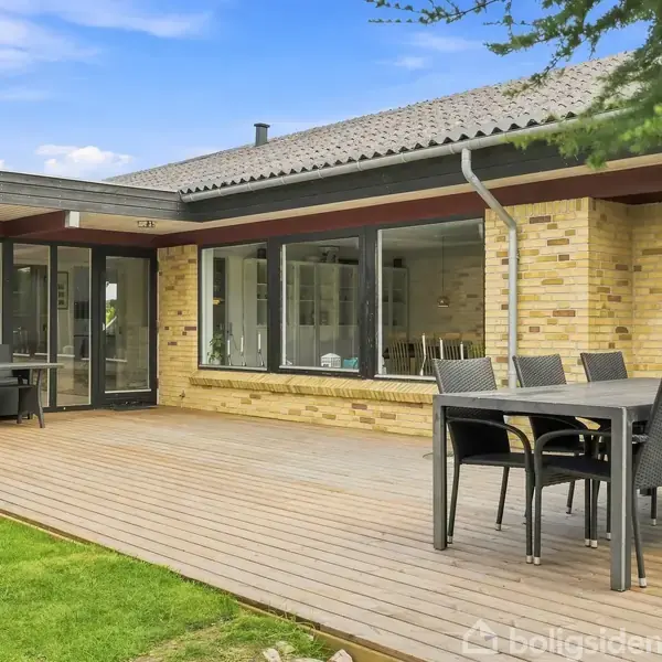 Terrasse med spisebord og stole står på trædæk foran et moderne hus med store vinduer og gul murstensfacade. Græsplæne og træer omgiver området under en klar blå himmel.