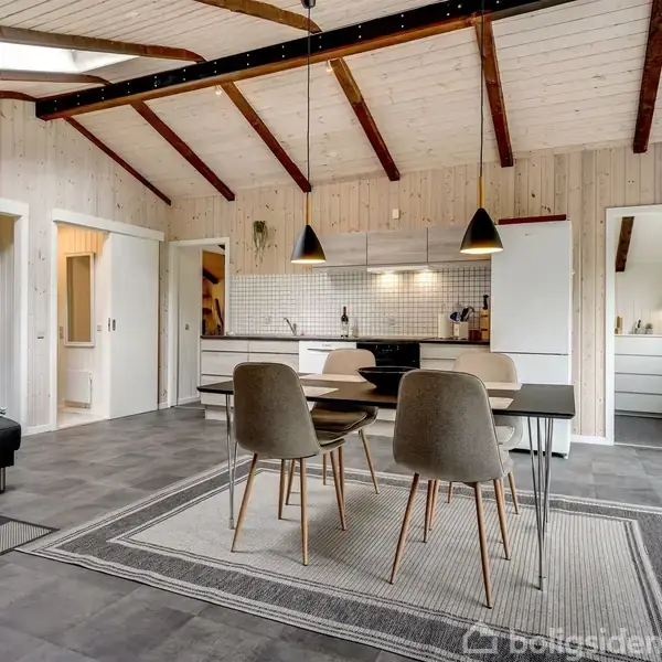 Et moderne køkken-alrum med spisebord og fire stole. Træloft med synlige bjælker over lyse trægulve. Flere døre fører til andre rum, og et stort vinduesparti slipper naturligt lys ind.