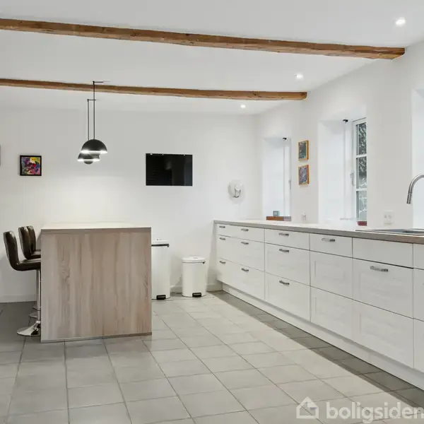 Et moderne køkken med hvide skabe, en lang bordplade og en central ø med barstole. Over øen hænger en sort lampe. Rummet har hvide vægge og flisegulv.