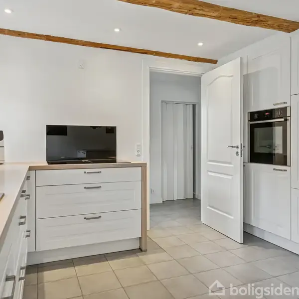 Hvide skabe og skuffer i et køkken, en åben dør ved siden af en indbygget ovn. En køkkenmaskine og en lille skærm står på en bordplade. Fliser på gulvet, træbjælker i loftet.