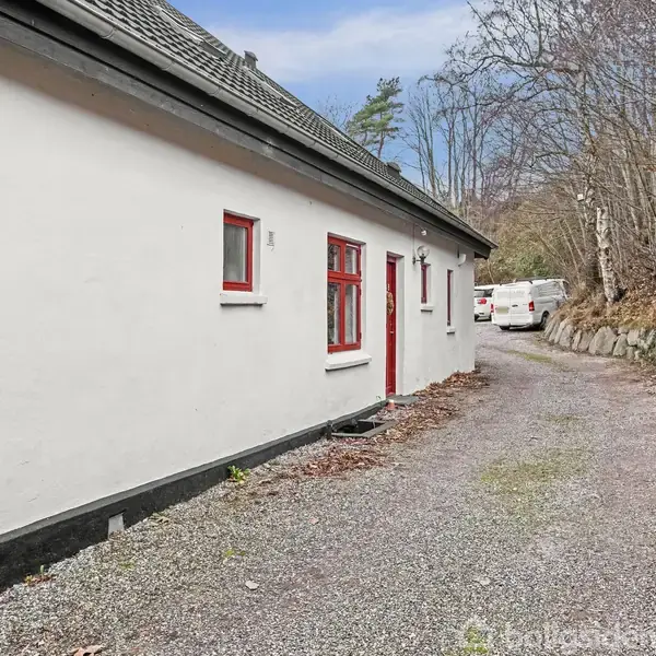 Hus med hvid facade og røde vinduer ligger langs en grusvej. Der er skov på den ene side og flere biler parkeret længere fremme.