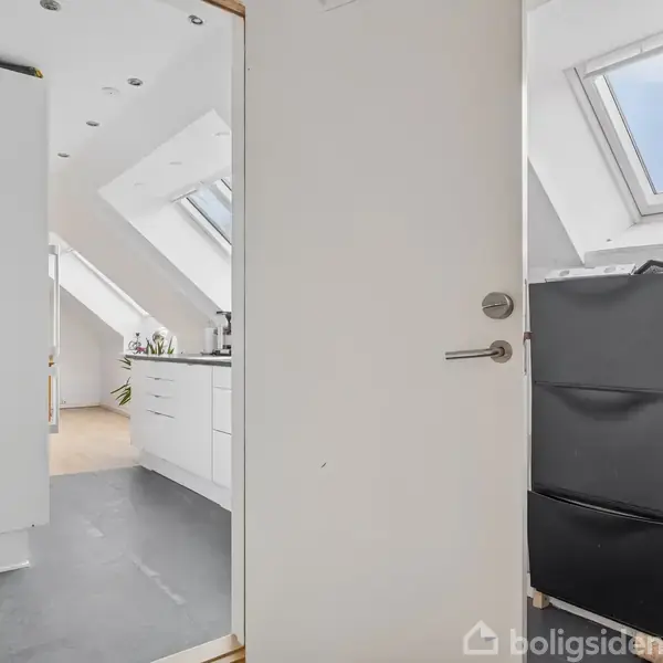 Dør åbner til et lyst køkken med skråvinduer, hvide skabe og sort skuffearrangement. Skabene er oplyst af naturligt lys fra vinduerne i loftet.