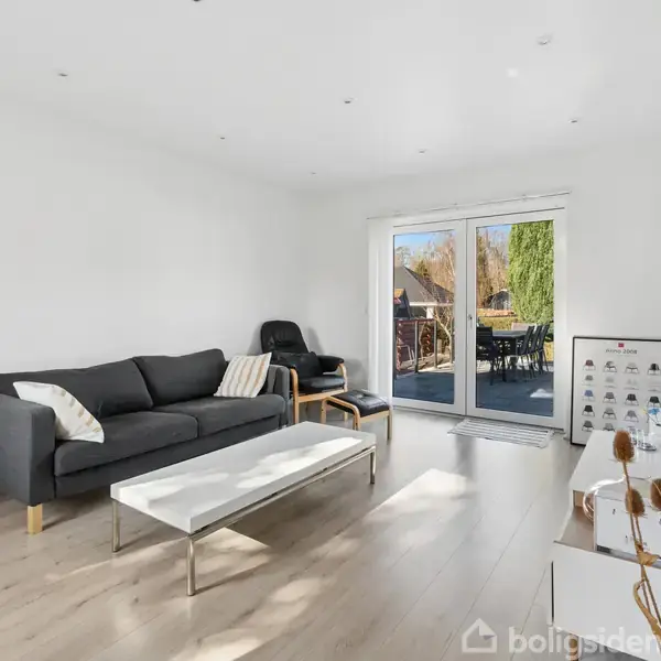 Sort sofa står i en lys, moderne stue med trægulv. Sofaen har puder og en lænestol ved siden, mens et stort vindue åbner ud til en terrasse med haveudsigt.