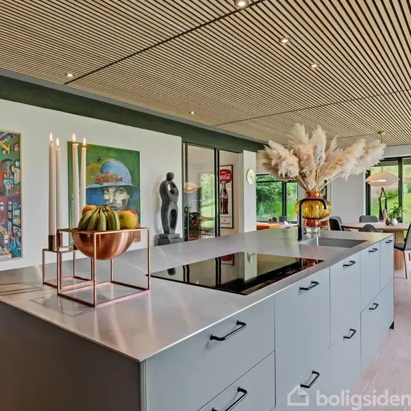 Køkkenø med induktionskogeplade og dekorationer som lys og græskar; i baggrunden ses moderne kunstværker på væggen. Store vinduer kaster lys over et stilfuldt indrettet rum.
