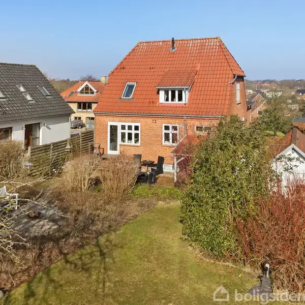 Et rødt murstenshus med en rød tag i tre etager står i en have med træer og buske. Nabohuse med lignende arkitektur ses i baggrunden under en blå himmel.