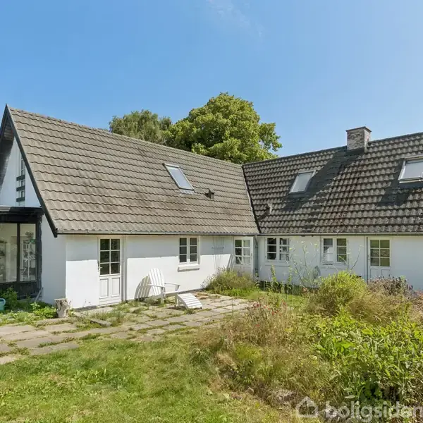 Hvidt hus med skråtag, placeret i en frodig have. Døre og vinduer er synlige. En pergola med glas er tilknyttet på venstre side. Blå himmel og solskin.