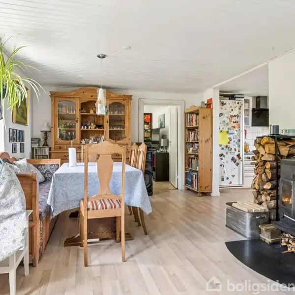 Et spisestue med et spisebord dækket af en dug; der står stole omkring det. En brændeovn står til højre med en gryde ovenpå. Ved siden af er en stabel brænde.