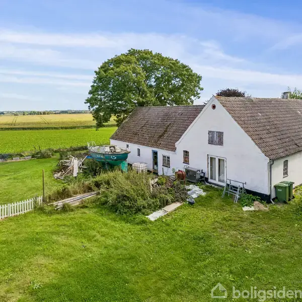 Hus med hvid facade og brune tage står på grøn græsplæne, omgivet af marker og træer under en klar blå himmel. En lille hvid havelåge ses ved siden af huset.