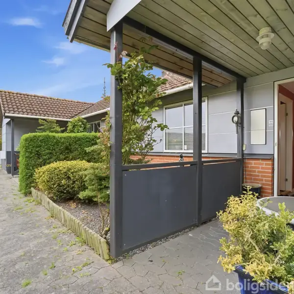 Radkejendom med overdækket veranda, hvor en stol og planter står. En åben dør fører ind, og fortovet løber langs huset omgivet af grønne hække under en blå himmel.