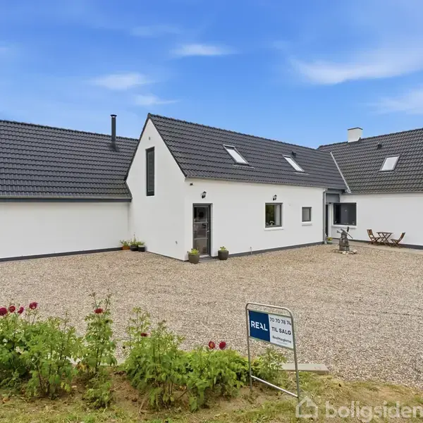 Et hvidt, moderne hus med sorte tagsten står på en grusbelagt gårdsplads. Der er et skilt foran med teksten "REAL Mæglerne Tilst Til Salg". Græsplæne og blomster omgiver huset.