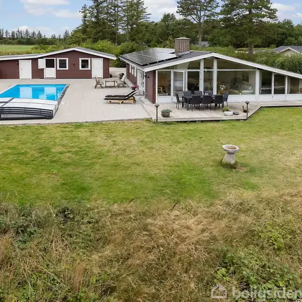 En moderne villa med store vinduer står ved siden af en udendørs pool og terrasse med havemøbler, omkranset af en grøn græsplæne og skov i baggrunden.