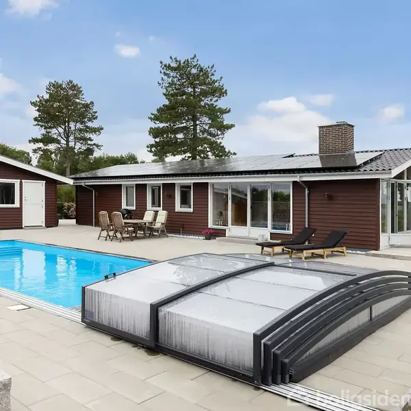Et træhus med store vinduer står ved siden af en blå udendørs pool. Omkring poolen er der en flisebelagt terrasse med havemøbler, omgivet af træer.