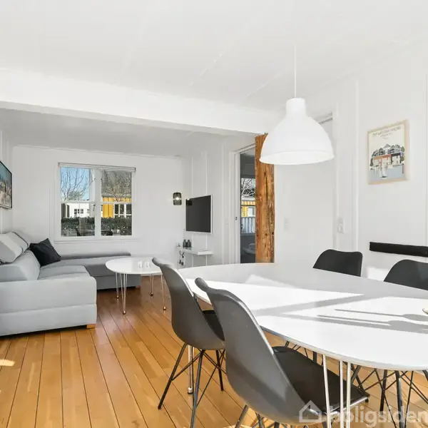 En moderne stue med træplankegulv, hvor en grå sofa står ved et vindue. Et rundt spisebord med sorte stole står under en hvid lampe. Væggene bærer kunstplakater.