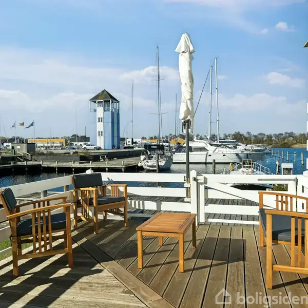 Terrasse med træmøbler og lukket parasol står ud mod en marina med flere både ved kaj. I baggrunden ses en hvid bygning og en blå himmel.