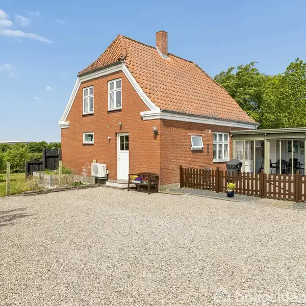 Rødt murstenshus står stille i en åben have omgivet af træer og grusbelagt gårdsplads. Terrasse med hvidt tag og brun stakit. Klart, solrigt vejr med blå himmel.