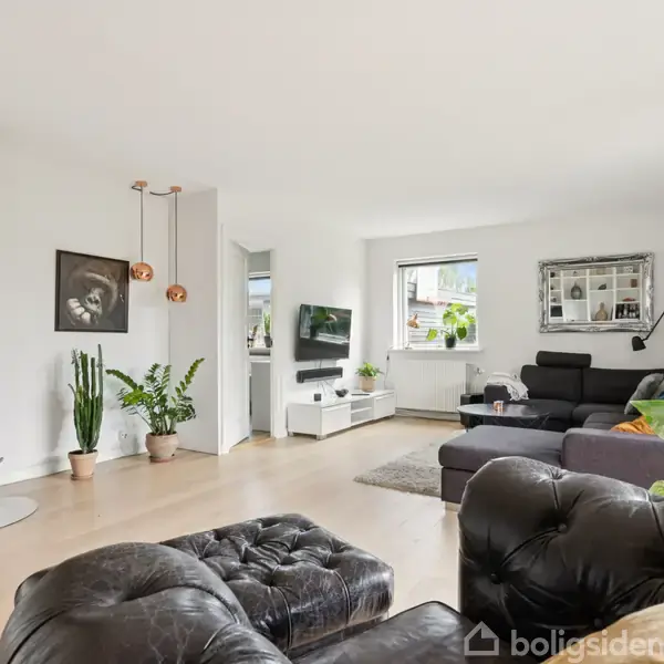 Brændeovn står i hjørnet og opvarmer rummeligt opholdsrum med træmøbler og moderne indretning. Store vinduer giver naturligt lys, mens planter tilføjer grønne elementer. Sorte sofaer og tv-møbel er centralt placeret.