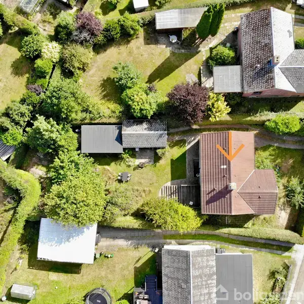 Hus set fra oven, omgivet af frodige træer og haver, med en brun tag og en hævet terrasse. Til højre ses en vej langs ejendommen.