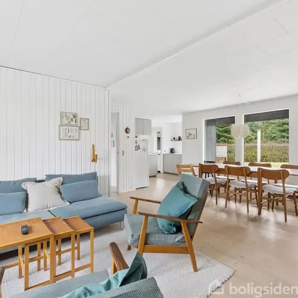 Sofa står i stue med træmøbler, vendt mod store vinduer, der viser en have. Køkkenbord med stole er i nærheden. Vægge er lyse, og rummet har en skandinavisk stil.