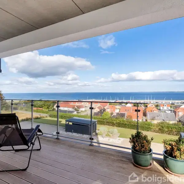 En balkon med et glasgelænder har to liggestole og to planter i krukker. Den har udsigt over huse med røde tage og havet under en delvist skyet himmel.