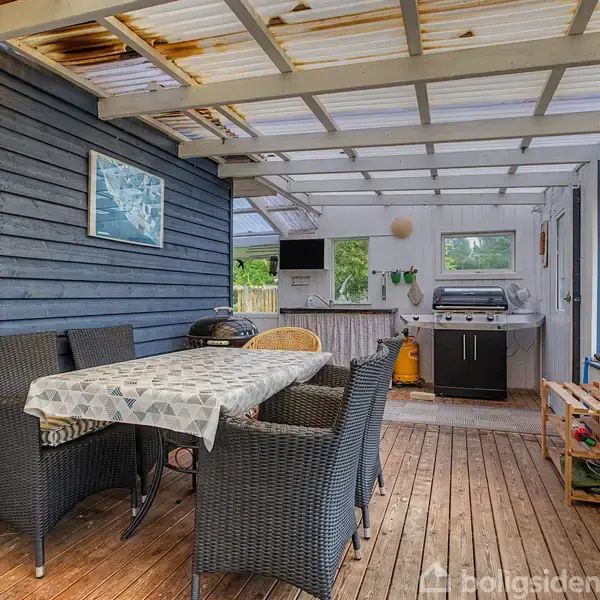 Et udendørs overdækket område med et spisebord og stole. En grill står tæt ved væggen. Tømrede vægge omringer området, og der er en kost på væggen.