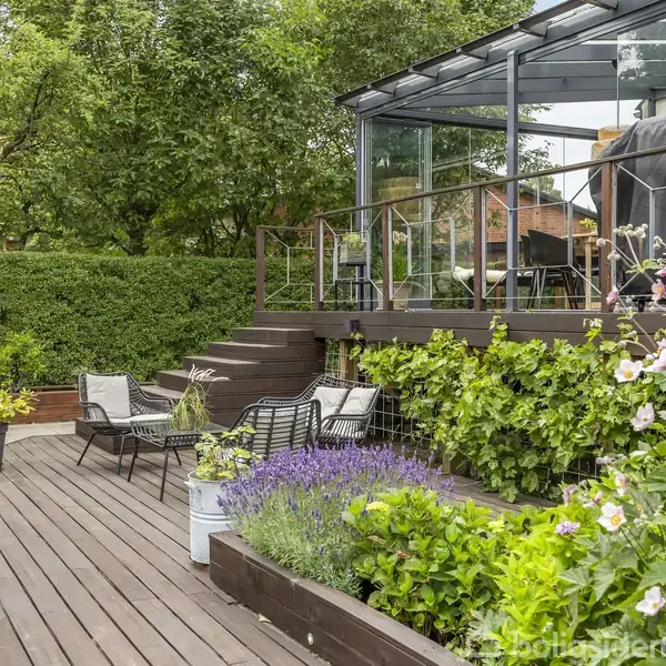 Terrasse med møblerede siddepladser omringet af grønt løv og blomster. En glasoverdækket udestue ligger hævet i baggrunden. Omgivelserne er frodige med hække og træer.