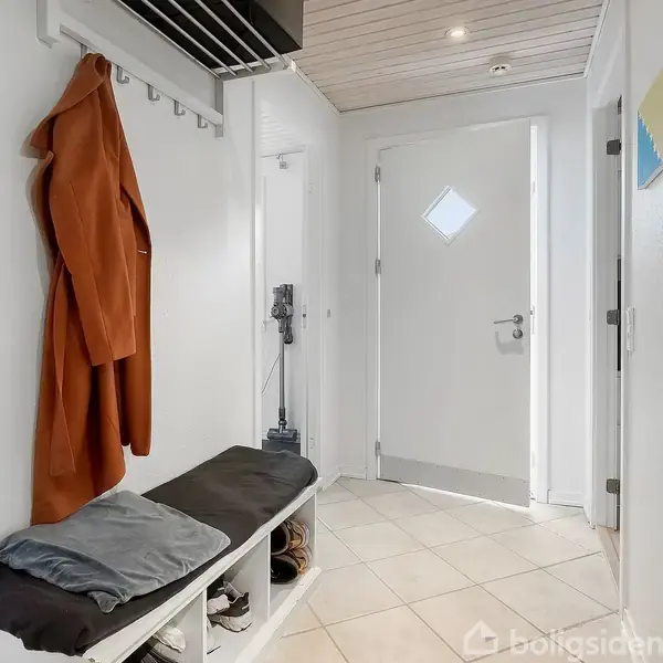 Frakke hænger på knagerække; skab ved siden af bænk med pude og sko, i entré med hvid dør og flisegulv.