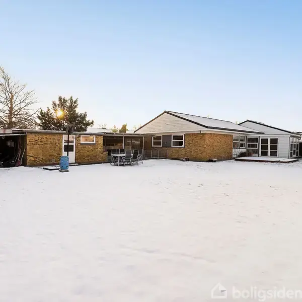En gulstensvilla står i en snedækket have. Der er et terrasseområde med havemøbler, mens tomme træer omkranser ejendommen under en lys, klar himmel.