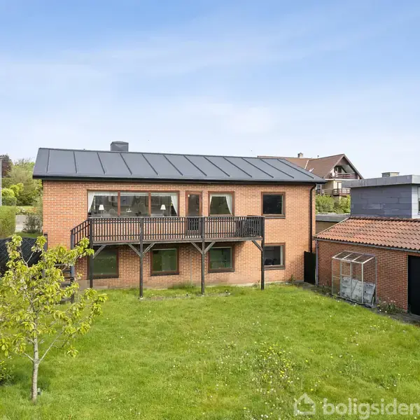 Rødt murstenshus med sort tag og balkon, beliggende i en grøn have med græs og træer. Nabohuse ses i baggrunden under en klar, blå himmel.