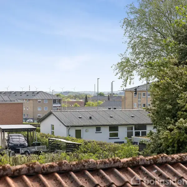 Huse med flade tage står i forgrunden omgivet af træer og haver. I baggrunden ses flere etagebygninger under en klar blå himmel.