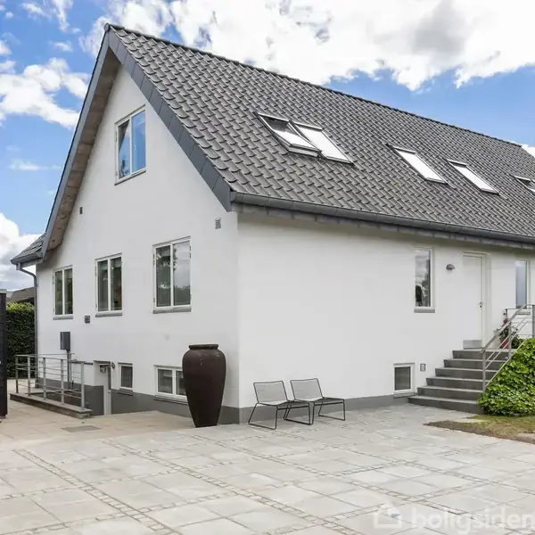 Hvid to-etagers hus med gråt tag står i et udendørs gårdareal. En trappe fører op til hoveddøren, mens møbler og en stor krukke pynter terrassen. Skyfri blå himmel.