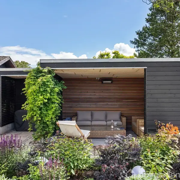En moderne havepavillon med et lysegråt loungesæt står på en terrasse omgivet af rige grønne planter og blomster, under en blå himmel med lette skyer.