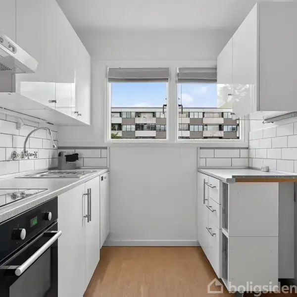 Et lyst, moderne køkken med hvide skabe og fliser. En ovn og kogeplader er indbygget. To vinduer giver udsigt til en boligblok. Trægulv og en bordplade med vask.