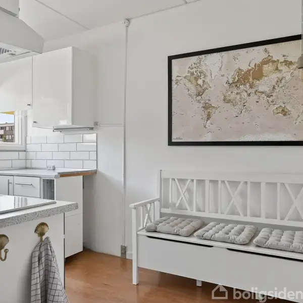 En hvid bænk med puder står i et lyst køkken, dekoreret med et kort på væggen. Rummet har hvide skabe, fliser og en moderne kogeplade.