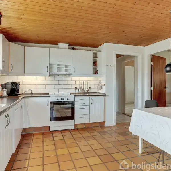 Køkken med hvide skabe og hvide fliser over en moderne komfur. Spisebord med hvid dug til højre, omgivet af sorte stole. Træloft og flisegulv skaber et varmt miljø.