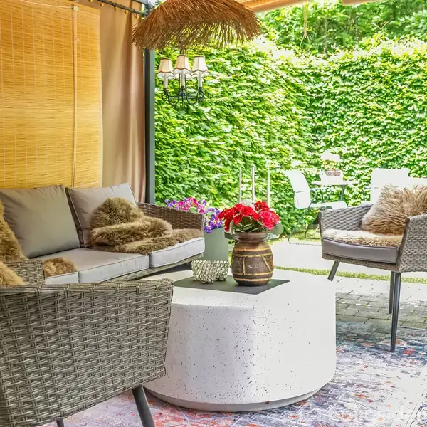 Et overdækket terrasseområde med flettede møbler, herunder sofa og stole, er pyntet med puder og tæpper omkring et rundt bord. Området er omgivet af frodige grønne hække.