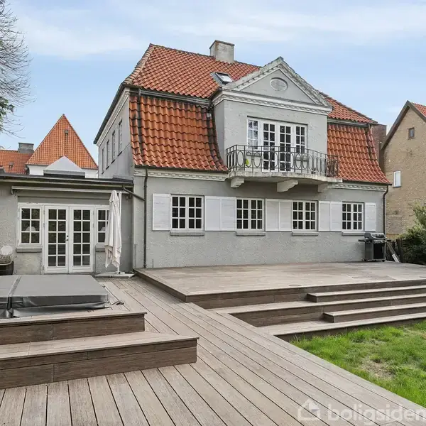 Et stort hus med rødt tegltag og kviste, omgivet af en rummelig træterrasse og en velplejet græsplæne. Huset har hvide vinduer og står mellem andre bygninger.