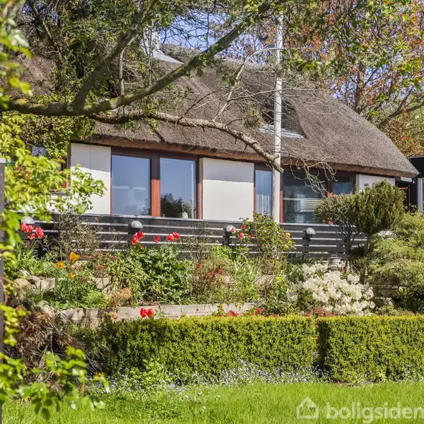 Stråtækt hus omgivet af frodig have med blomster og buske. Sollys skinner gennem træernes grene, der også giver skygge til huset. Atmosfæren er fredfyldt og idyllisk.