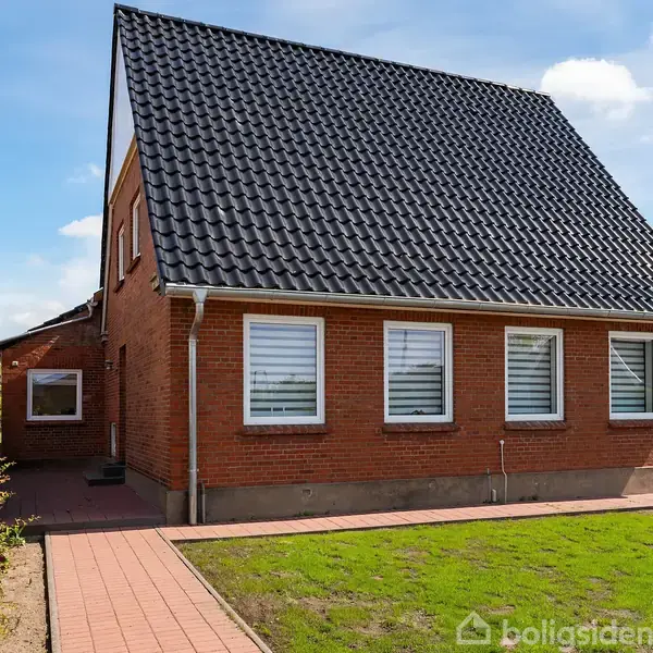 Huset har en rød murstensfacade med fire hvide vinduer. Det sorte tag er stejlt. Omgivet af en velholdt græsplæne og en flisebelagt gangsti. Skyfri himmel i baggrunden.