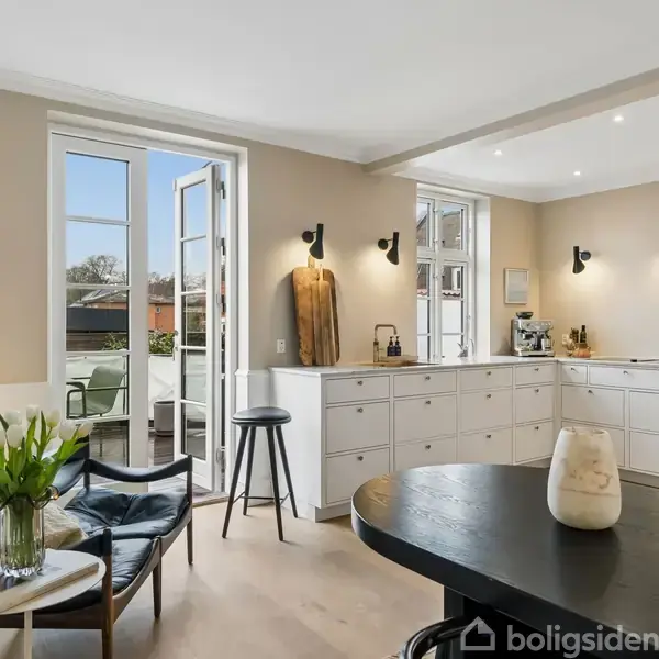 En moderne stue med møbler og køkken, set i et lyst, skandinavisk design. Der er en sofa, et rundt bord med en lysestage, køkkenskabe, og en åben fransk altandør.