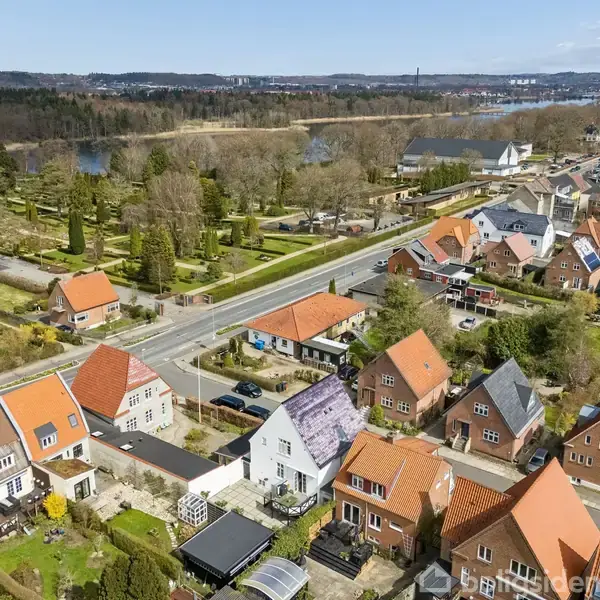 Række af huse med røde tage står langs en vej. Bagved ligger en park med træer og en sø i baggrunden under en blå himmel.