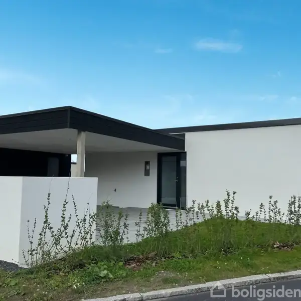 En moderne, hvid, en-etagers villa med fladt tag står i en forstadslignende have. Der er en tilbygget carport på venstre side med flisebelagt indkørsel og spredt vegetation omkring.