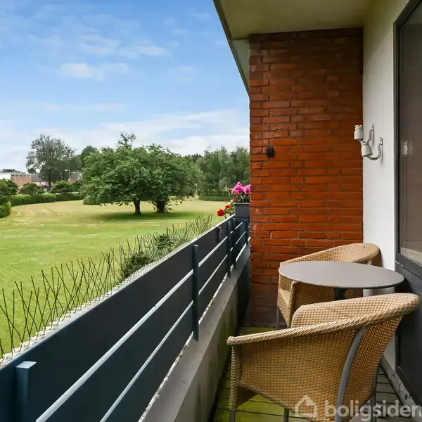 Balkon med to flettede stole og et rundt bord. Udsigt over en grøn have med træer og buske. En murstensvæg og et vindue er synlige til venstre.