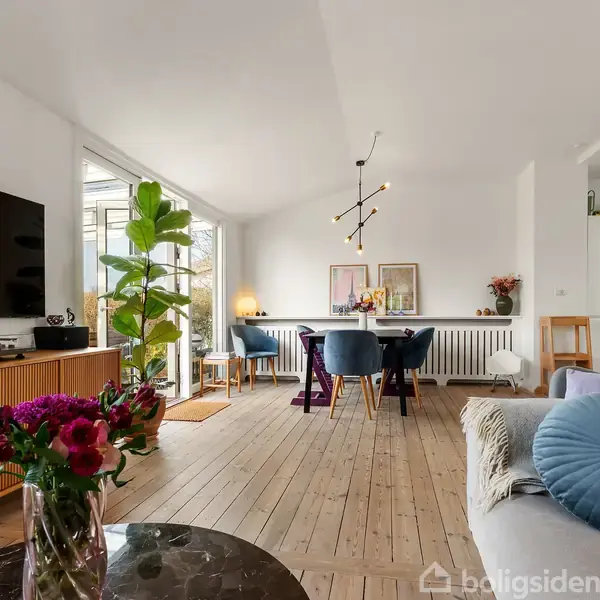 En stue med trægulv har en sofa med puder, et marmorbord med blomster, en stor plante ved terrassedøren og en spisestue med bord, stole og kunst på væggen.
