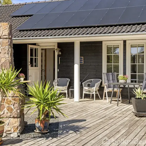 Solpaneler er monteret på taget af et sortmalet hus. På træterrassen står havemøbler og plantekrukker med grønne planter. Dørene åbner ud mod terrassen, skaber en indbydende udendørs atmosfære.